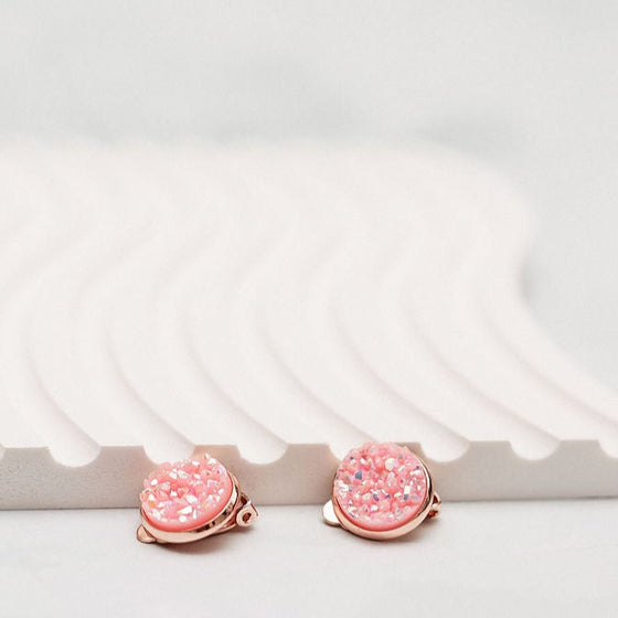 "Two pink sparkly faux druzy earrings set in rose gold clip-on style. One showcases the front with shimmering pink faux druzy gems, while the other displays the back clip-on mechanism. Both earrings elegantly laid on an off-white wavy plate background."