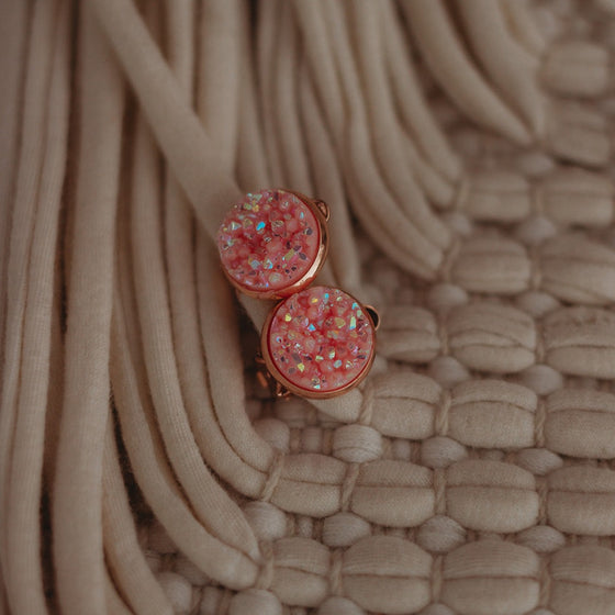 "Pink sparkly faux druzy earrings set in rose gold, resting on a textured woven basket surface, casting graceful shadows.