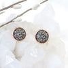 "Silver druzy earrings in a rose gold setting, elegantly placed on a wood floor with a backdrop of shimmering white crystals."