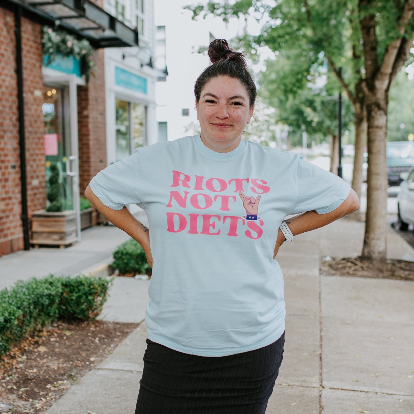 Person wearing a light blue t-shirt with 'Riots Not Diets' text on a sidewalk.