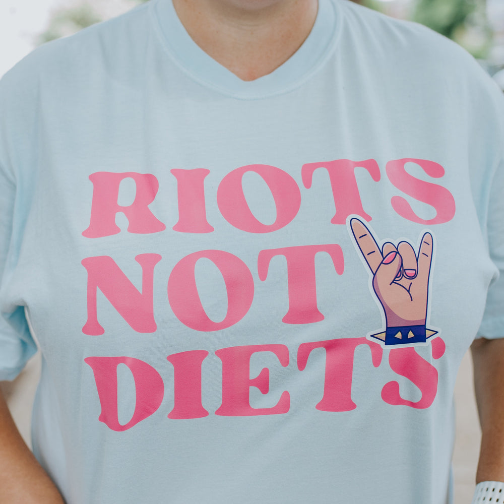 Person wearing a light blue t-shirt with pink text 'RIOTS NOT DIETS' and a hand sign.