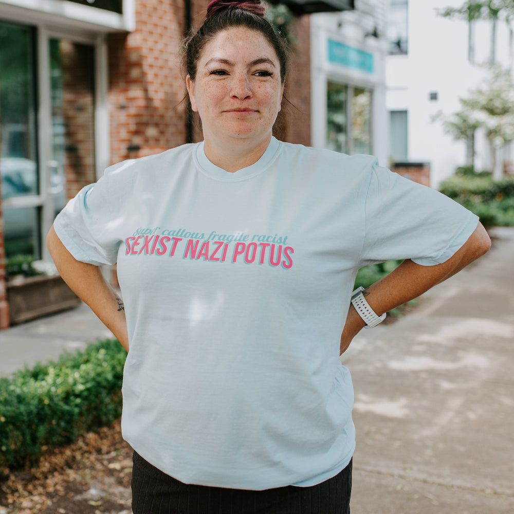 
                  
                    Person wearing a light blue t-shirt with text, standing on a sidewalk.
                  
                