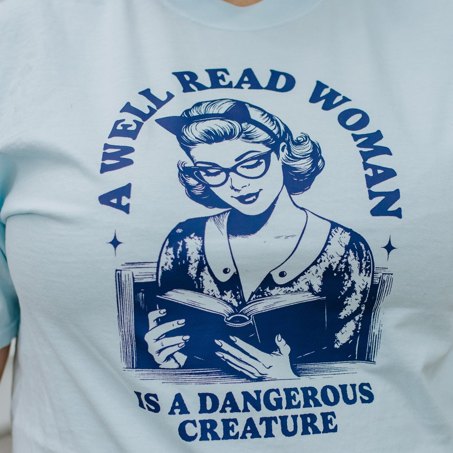 Light blue t-shirt with a graphic of a woman reading a book and text 'A Well Read Woman is a Dangerous Creature' in blue.