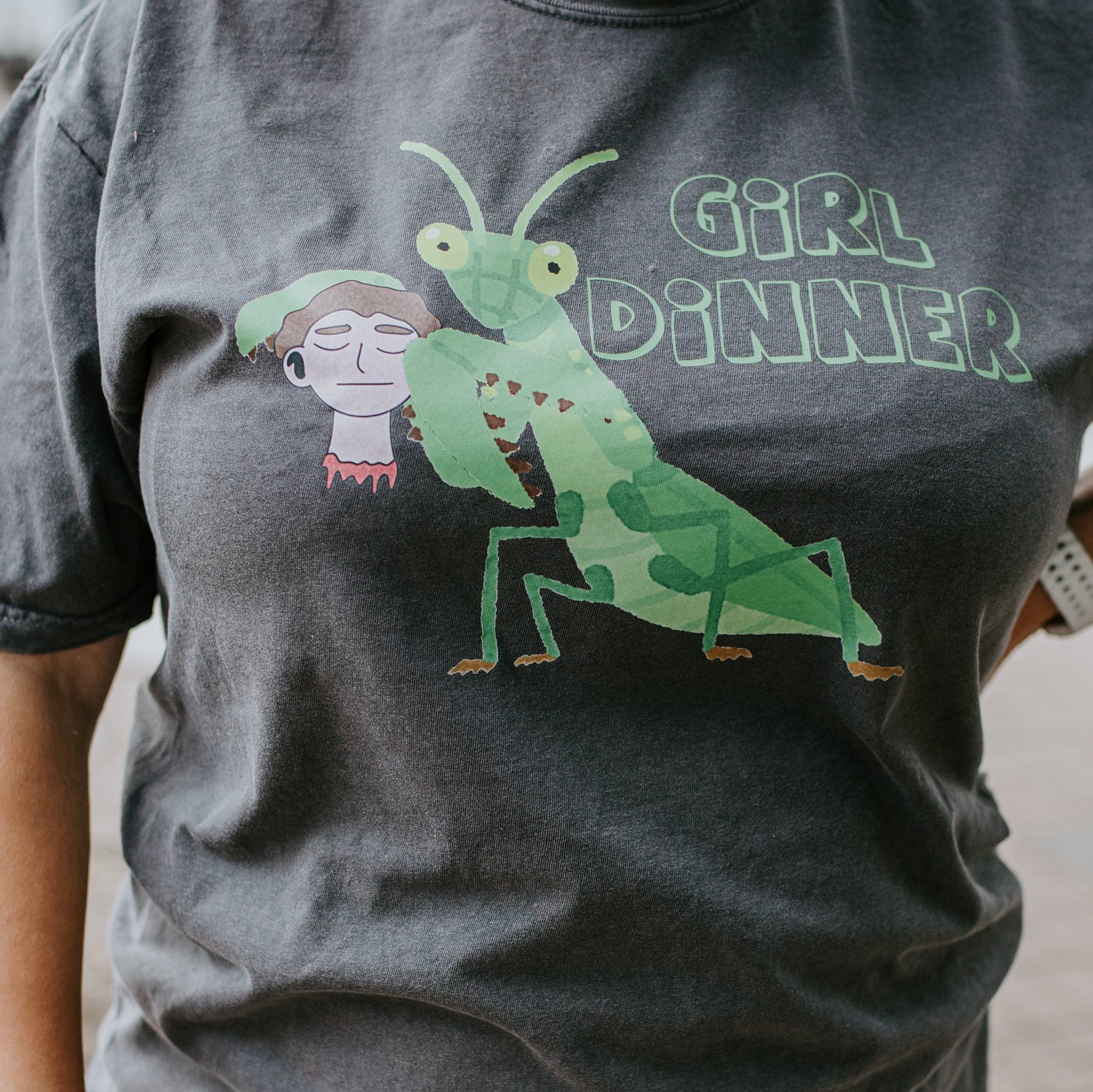 Person wearing a t-shirt with a graphic design of a green praying mantis and a person, with text 'Girl Dinner' on a dark gray background.
