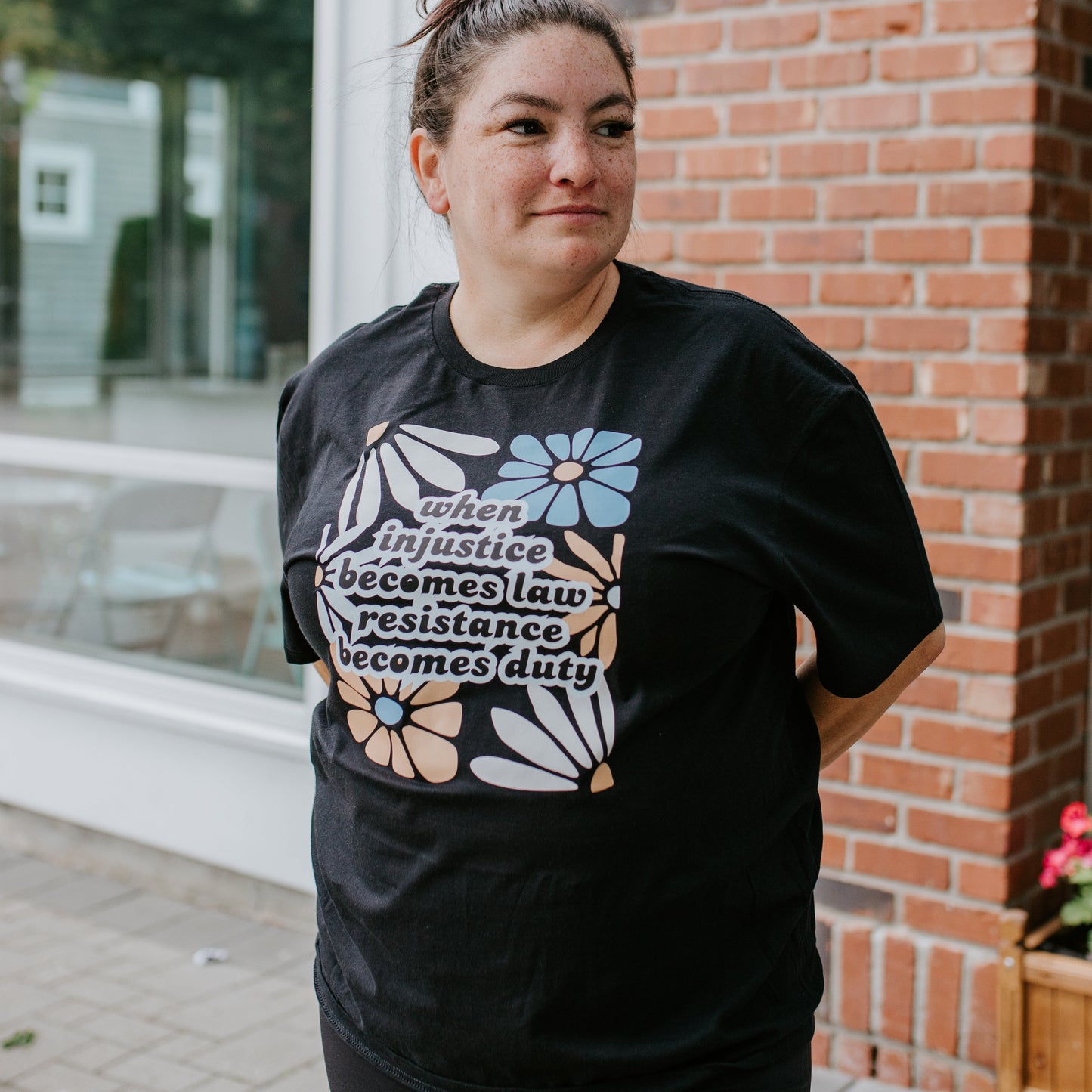 Person wearing a black t-shirt with a graphic design standing outdoors next to a brick building.