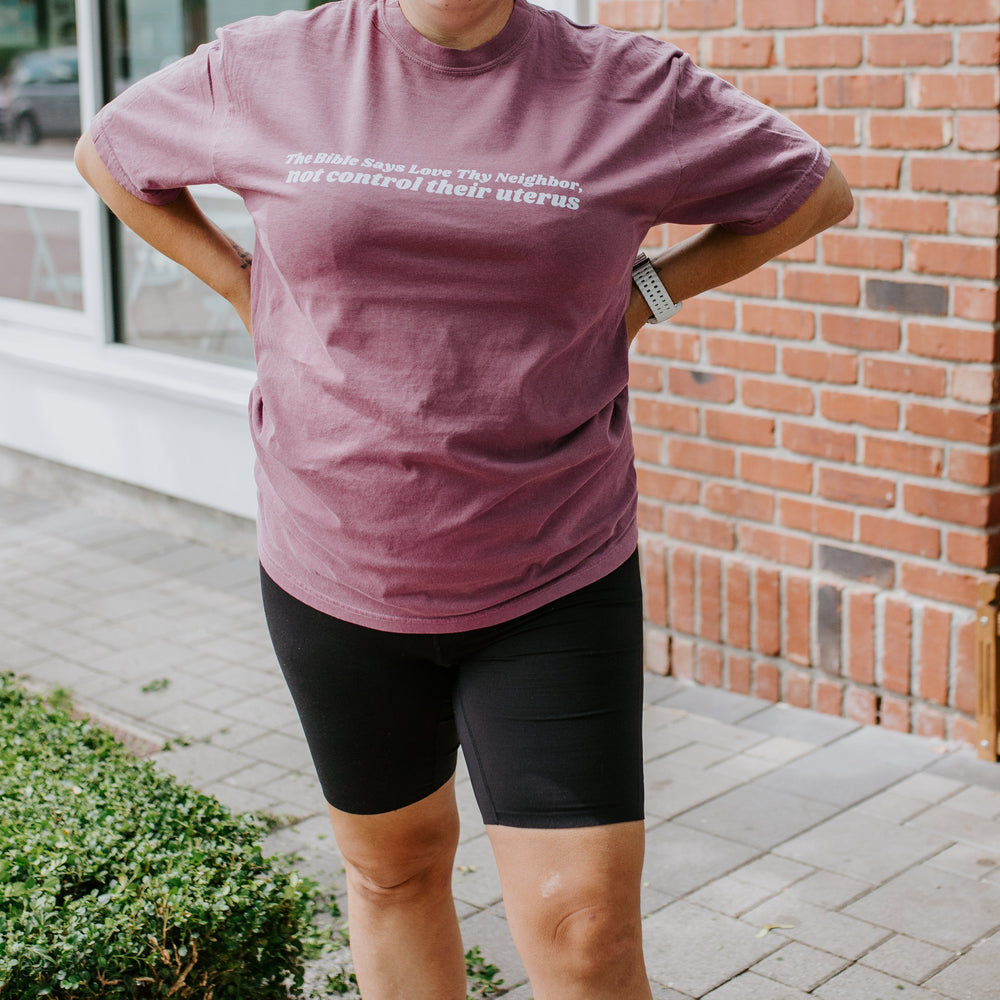 
                  
                    Person wearing a purple t-shirt with text, standing outside a building with a brick wall.
                  
                