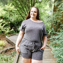  "Woman with dark hair wearing a dark grey shorts romper with a drawstring, standing on a forest pathway with trees in the background.